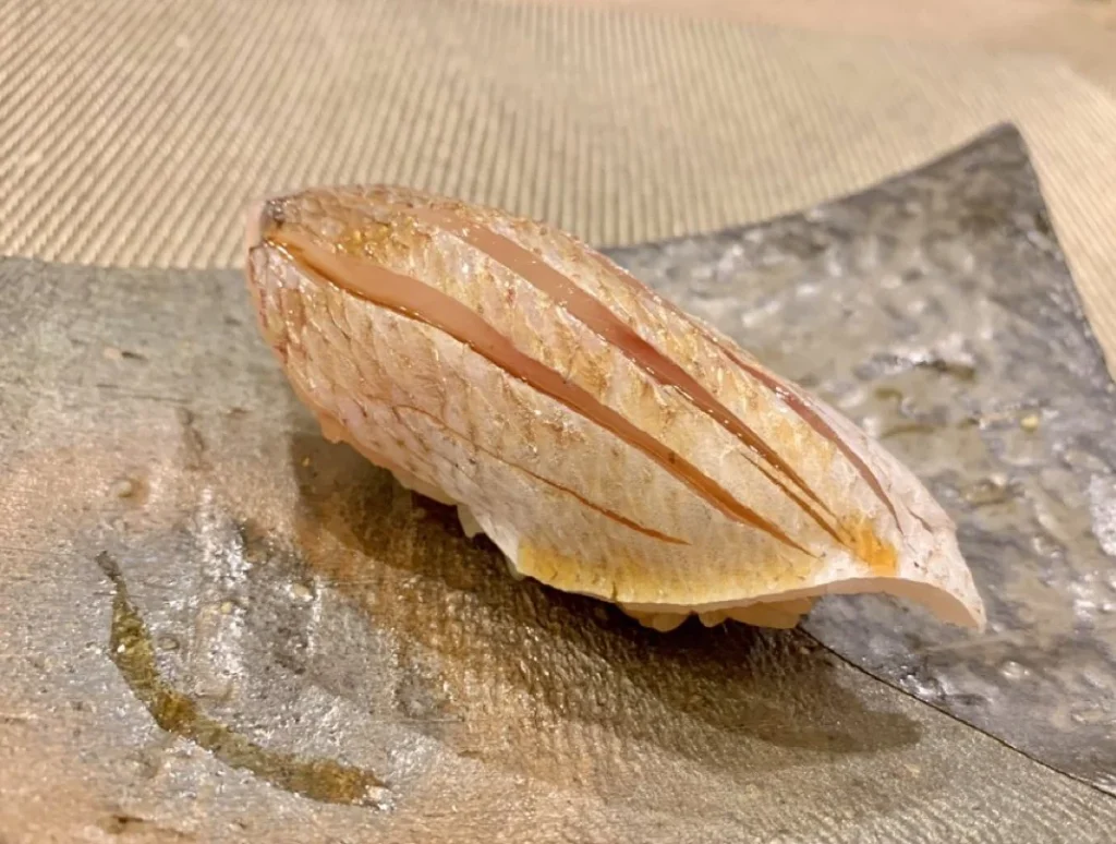 Kasugo Nigiri Sushi Single piece of kasugo nigiri with scored silvery pink skin on a ceramic plate