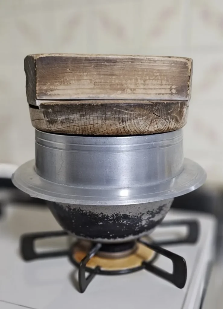 Traditional metal hagama with a thick wooden lid heating over a gas stove`
- メディアタイトル：`Traditional Hagama with Wooden Lid