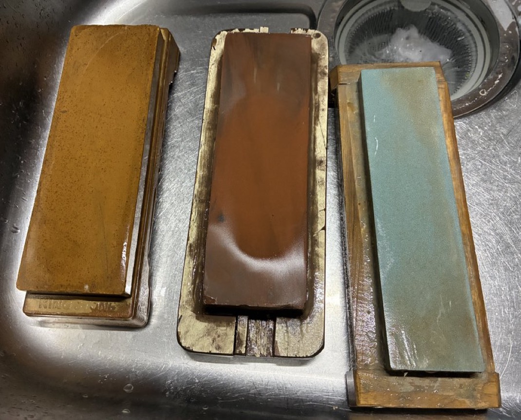 Three wet Japanese whetstones resting on holders beside a kitchen sink