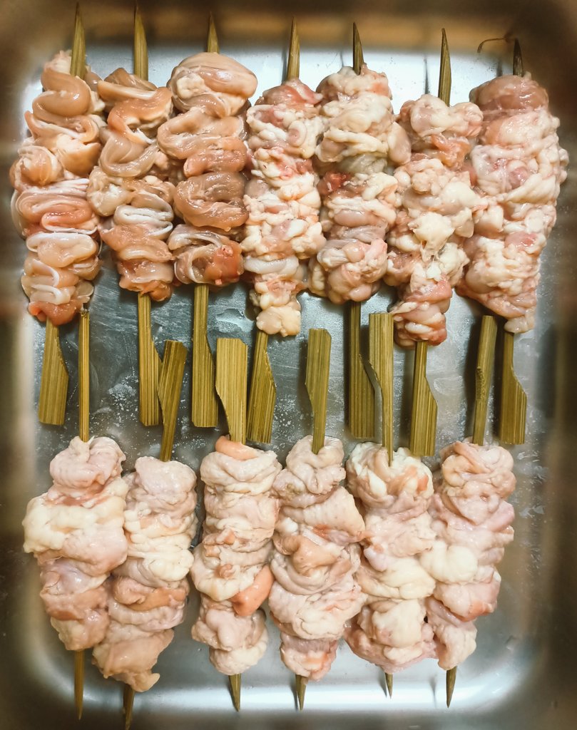 Parboiled chicken skin folded tightly onto flat bamboo skewers before grilling