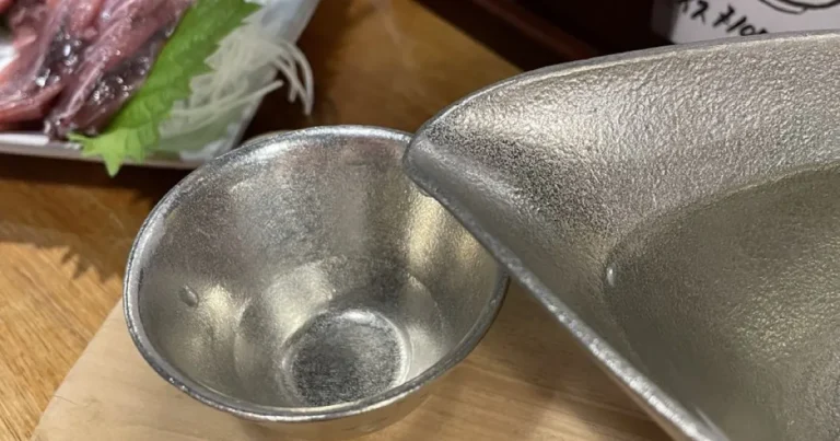 Silver-toned sake cup and pouring vessel beside sliced fish at a Japanese table