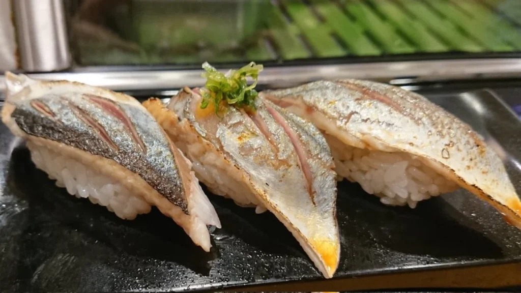 Sanma Nigiri Sushi Three pieces of silver-skinned sanma nigiri arranged on a black plate at a sushi counter